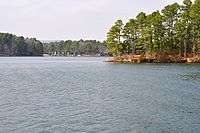 The blue waters of Lake Catherine with a tall pine tree-covered point jutting out into it and camp facilities visible on the shore in the background