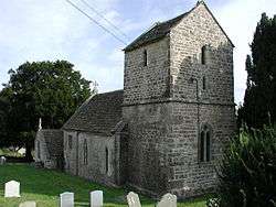 Gray building with tower at the near end. Trees to right. Gravestones in front