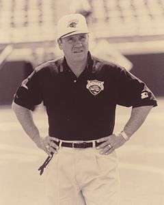 Candid sepia-toned photograph of Pasquale wearing a black polo-style shirt and white baseball cap both bearing the logo of the Jacksonville Jaguars and standing on a football field with arms akimbo