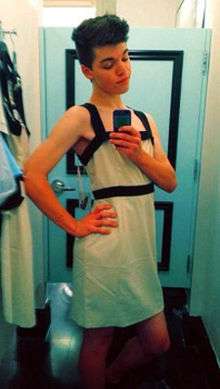 Teenaged, white, transgirl with dark hair wearing a white dress and posing in front of a mirror, taking a photograph of herself using a camera phone.