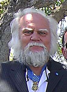 Head and shoulders of a white man with wild white hair and a full beard wearing a dark suit with a star-shaped medal hanging from a blue ribbon around his neck.