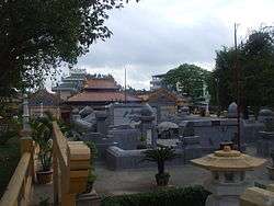 A cemetery with densely packed headstones made of cement. Almost no grass or flora can be seen. Many of the headstones are elaborate with a yellow or orange-tiled roof in the manner of an East Asian temple.