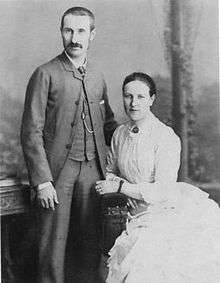 photo of man dressed in suit standing with woman in white dress seated beside him