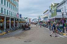 A street, surrounded on both sides by two- and three-story buildings. One person is riding on horseback in the middle of the street, while others are walking on the sidewalk. Bikes are parked at the curb.