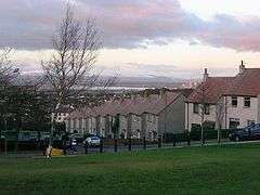 A row of housing on a downhill street with bare trees lining the opposite side, a view of the Forth Valley and Ochil Hills in the background.