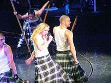 Madonna wearing a white T-shirt and a black-and-white patterned skirt, holding a microphone. She is surrounded by dancers in similar attire, carrying long poles.