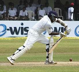 A man standing on a cricket pitch, wearing a white cricket kit and blue helmet leans forward to play the ball with a bat held out in front. The seating area of the ground is visible in the background.