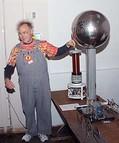  A man wearing coveralls with his left hand near the metal sphere of a Van de Graaff generator. His hair is standing on end due to electrostatic repulsion.