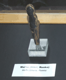 brown female figurine on small, square transparent base with id card that says in English and in Czech, "Mal'ta (Siberia, Russia)"