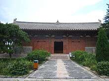 A Chinese temple building that is red. In front of it is a pathway and trees and flowers.