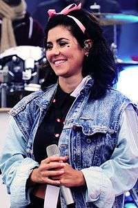 A young brunette woman, standing in front of a drumkit, holding a microphone and smiling. She is wearing a denim jacket, has a pea-sized heart-shaped mark painted on her left cheek and has a pink bow in her hair.