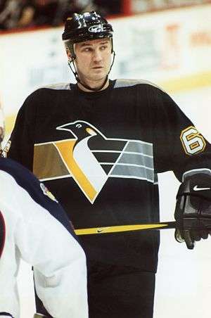 Hockey player in dark uniform. He holds his stick with both hands and looks onward, head tilted slightly.