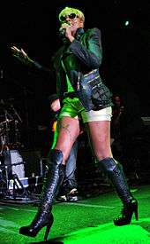 A woman walks while she sings. She wears sunglasses, a leather jacket and boots, and a gray blouse and short pants.