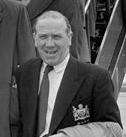 A black-and-white upper body photograph of a bald man. He is wearing a black suit with a tie.