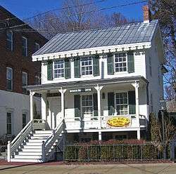 A two-story white wooden house with decorative touches, a pointed metal roof and green shutters. A sign in the center of the porch reads "Irvington History Center" while another at the right on the story below says "Frocks, Frills and Furbelous 1760-2009"