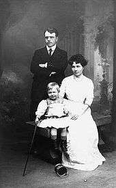 A studio portrait. A young man stands with his arms folded; he has dark hair, and is wearing a dark Edwardian suit, a white shirt with rounded collars, and a dark tie, To his right, a young woman sits on a wooden bench; she has dark, medium length hair, and is wearing a white blouse and a long white skirt. She holds a young fair-haired boy, who is wearing a light tunic with flared skirt and embroidery at the neck, dark boots and short socks. He holds a walking stick in his right hand. An empty paint tin lies on its side near his feet. The background has a colonnade and clouds in the classical romantic style.