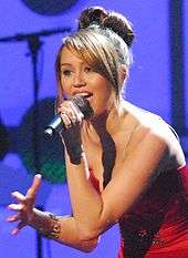 A three-quarter view of a female teen who faces left and leans slightly forward while singing into a black microphone. She wars a red ball gown and her straight brown hair is styled in a large bun.