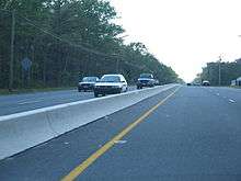 A four-lane road in a wooded area with a small concrete barrier in the center. Power lines are on either side of the road.