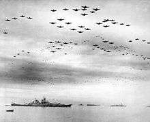 Black and white photo depicting a large number of aircraft flying in formation above a group of World War II-era warships