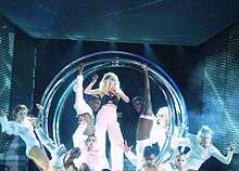 Gaga standing inside circular metallic rings in a white dress, surrounded by her dancers onstage.