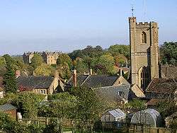 View of the roofs of houses with a prominent square church tower, interspersed with trees.