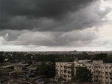 Daytime view over a city: de-laminating concrete housing blocks surrounded by rough slum tenements. In the middle distance, an expanse of trees: perhaps a park. Near the horizon, the largely concrete structures that compose the city continue. The whole is enveloped by an ominous sky filled with storm clouds promising imminent heavy rain.