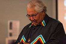 Photo of Justic Murrsay Sinclair taken during opening keynote address. He is seen, while looking down and smiling, wearing a black top with multi-coloured accents.