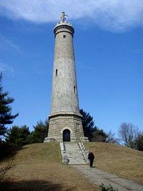 A very tall stone tower with a statue of a man at the top. It stands on a steep, grassy hill.  A pathway and stairs lead to a door on the front of the tower.  A person is walking along the pathway away from the tower.