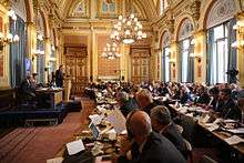 A large baroque yellow and gold room with a stage on the left and long tables filled with men and women in suits on the right.