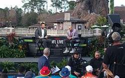 Two men sitting on director's chairs on a stage, listening to a question from an audience member