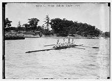 Four men sitting in a long canoe, in front of a wooded camp