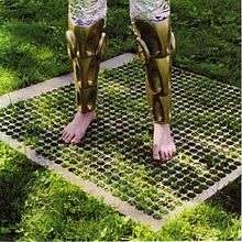 Two disembodied legs—barefoot with futuristic armor around the calves and wearing patterned tights—stand atop a grate placed atop green grass, partially covered by shadow