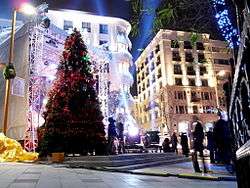 A large tree decorated under the night sky in red and green and surrounded by spotlights, city lights, and mid-rise buildings