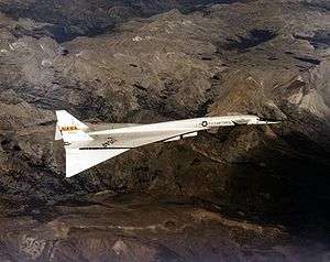 White delta-wing aircraft overflying mountains. The front of the fuselage features canard wings, and the wing tips are dropped.