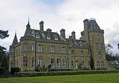 An imposing yellow-stone house in two storeys with attics seen from a slight angle. On the left are two circular turrets with conical roofs; on the right is a larger square tower with a truncated pyramidal roof. Rising from the roofs are dormers and chimneys.