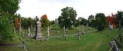 A panoramic view of the cemetery; a dirt road pass in front of the view from right to left. At center is a grey church-like structure, surrounded on all sides by various sized grave stones.