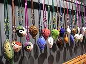 Colourful four-hole English pendant ocarinas hanging from a rail by elaborately-knotted 2- and 3-colour lanyards. Three are shaped like squat owls; several have low-relief or painted patterns.