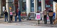 A group of seven people holding hand-lettered cardboard signs along a city street. The largest says "We are the 99%"