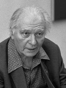 An elderly, balding man with swept back hair, wearing a suit. He faces the camera.