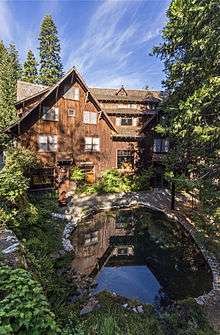 A rustic multi-story hotel sheathed in tree bark rests in a forest behind a reflecting pool.