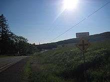 A winding country road with green trees growing on the right and a farm on the left. A white sign with blue lettering atop an intersection marker reads, "VILLAGE OF ORSON."