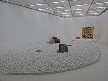 An exhibition gallery, with white pebbles on the floor, five images on slabs and a small block of sandstone to the back.