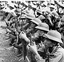 A large number of well equipped Asian soldiers sitting in rows on the ground, wearing pith helmets and holding assault rifles.