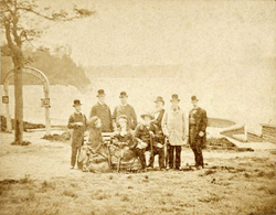 Photograph of three people, two women and a bearded man, seated on a park bench with six other men standing in back and waterfalls in the distance