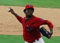 A right-handed man in a red baseball jersey and red baseball cap throws a baseball. His jersey and cap have a teal-colored shark on the front, and he is wearing a black baseball glove on his left hand.