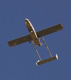 A large airborne machine photographed in flight from the ground looking up. The machine is pointed toward the top left side of the photo. Large wings can be seen protruding from the vehicle, along with the tail fin and metal peinces that attach it to the body of the aircraft. Visible in the machine's underbelly are a camera and landing gear, tail hook, and a blur in the back where a small propeller responsible for powering the machine can be found.