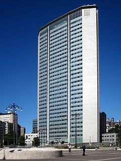 A narrow, unornamented skyscraper with blue-green glass windows in the middle and a tapered metallic skin on the sides rising above some trees at ground level against a blue sky