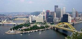  A park with a fountain between two rivers that meet. Many skyscrapers are behind, and several bridges cross the rivers.