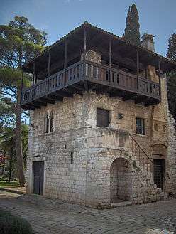 A small three-storey stone house with an exterior stone staircase to the first floor, and a wooden balcony around the upper floor