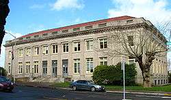 Photograph of the Astoria post office, a broad, institutional, two-story building in concrete or stone with a low-profile, red roof